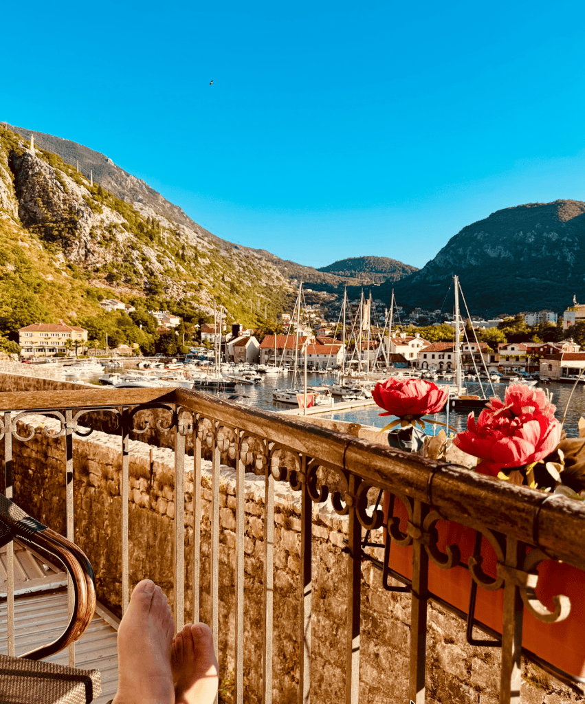 A view from a balcony overlooking a marina with boats, surrounded by mountains and colorful buildings, with a pair of feet resting on the railing and flowers in the foreground.