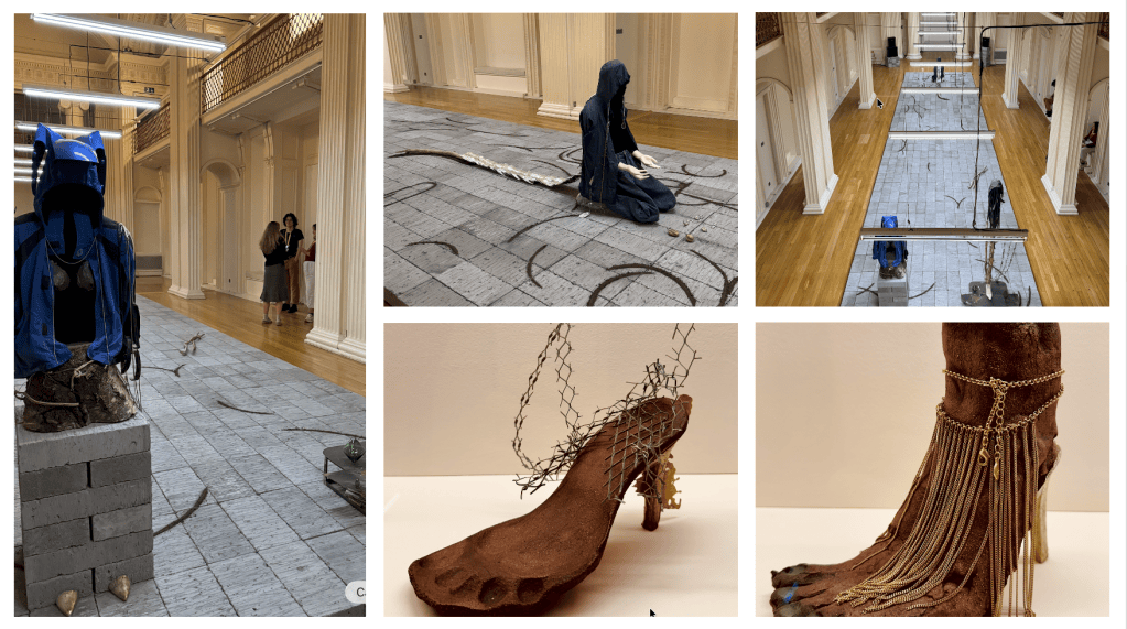 An art installation featuring a figure dressed in a blue cloak, positioned on a grey floor with abstract markings and a background of a spacious gallery.